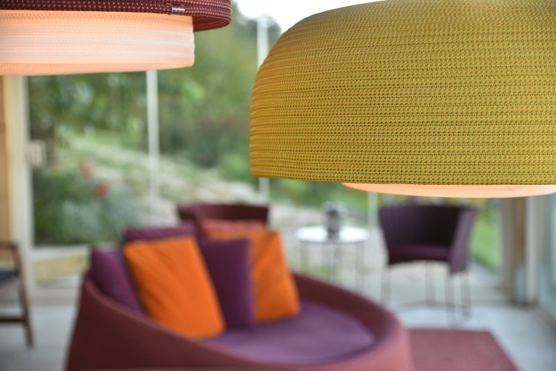 Grüne hängende Lampe im Vordergrund, im Hintergrund bunte Pölster und eine violette Couch