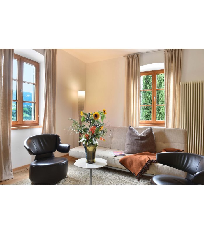 Helles Wohnzimmer mit kleinem weißen Tisch und frischen Blumen, umgeben von beiger Couch und zwei schwarzen Sesseln