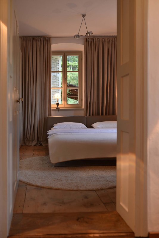 Blick ins Schlafzimmer mit gemütlichem Bett und weißer Bettwäsche, dahinter beige Vorhänge vor großem Fenster