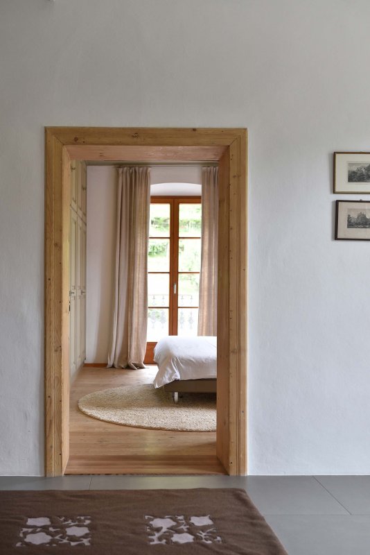Blick durch eine Holztür auf ein helles Schlafzimmer mit großen Fenstern und beigen Vorhängen