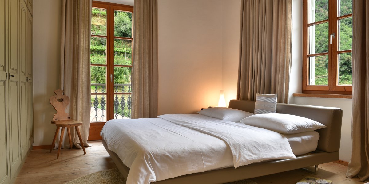 Gemütliches Bett mit weißer Bettwäsche vor großen Fenstern mit Blick ins Grüne