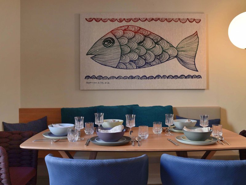 Gedeckter Tisch, blaue Stühle und dahinter ein Bild mit einem gemalten Fisch an der Wand